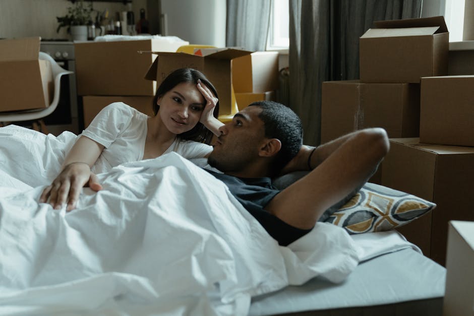 A young woman with long hair is lying in bed inside a room filled with cardboard moving boxes of various sizes, some stacked on a window sill and others arranged on the floor and around the bed. She a