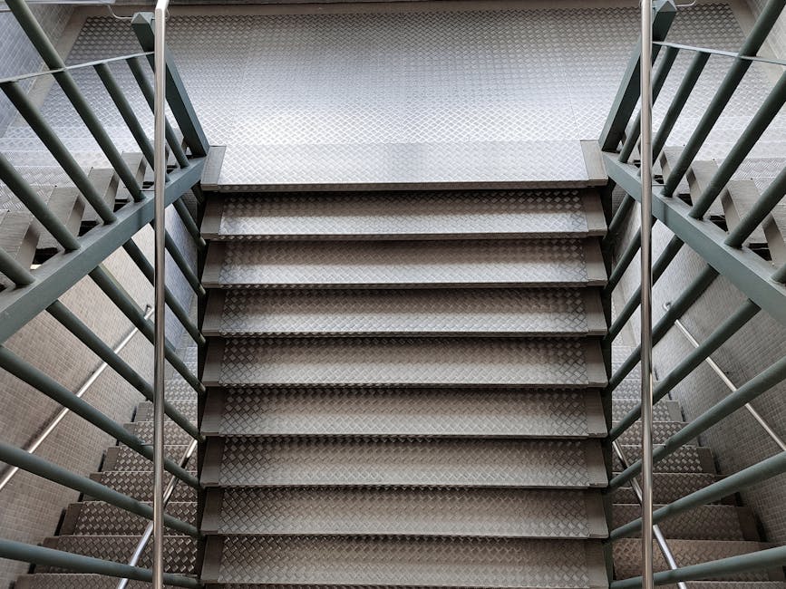 A high-angle view of a modern, multi-level staircase inside a building, with black metal railings and yellow safety strips along the edges of each step. The staircase features a curved section at the 