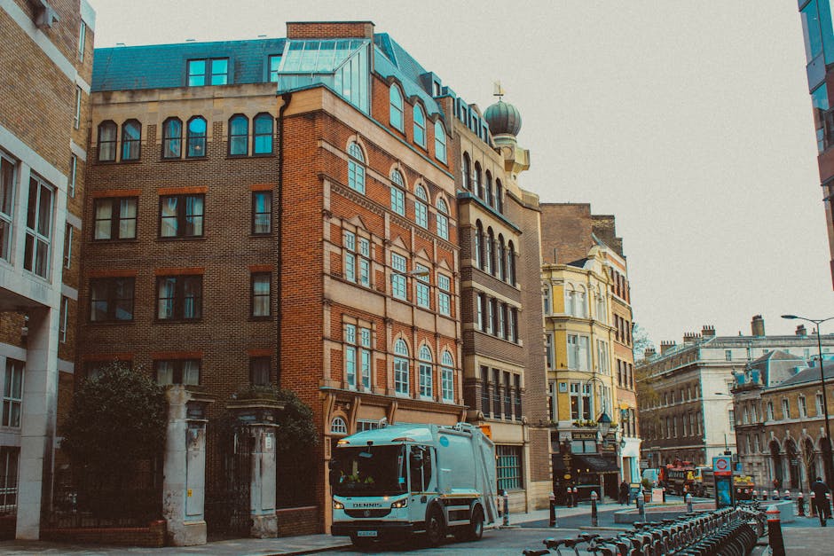 A white Iveco moving van parked on a narrow urban street with tall, multi-storey residential buildings on either side. The van is positioned centrally on the pavement, partially blocking the path. The