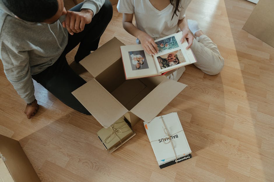 An overhead view of two individuals during a home relocation, with one person sitting on the wooden floor and the other kneeling beside a partially open cardboard box. Inside the box, several printed