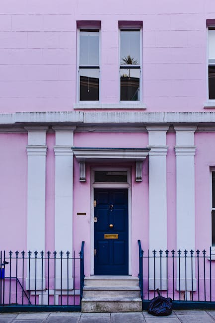 A close-up view of a pink residential building's exterior featuring a central dark blue front door with a brass mail slot and door knocker, accessed via a short set of concrete steps with metal handra